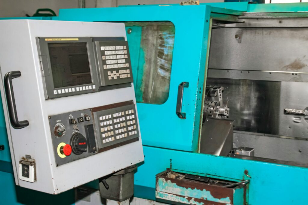 CNC machine control panel with screen, keypad, and emergency stop on a gray cabinet beside teal industrial equipment.